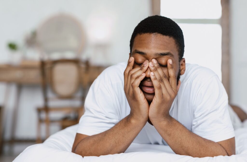 A young man waking up with irritated eyes, rubbing them while he lays on a white bed, propped up on his elbows.